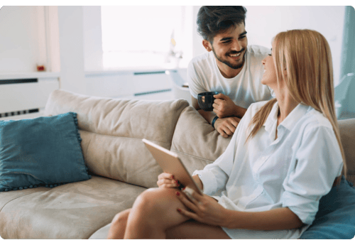 Man en vrouw zittend in een bank, lachend naar elkaar; de vrouw houdt een tablet vast en de man een kop koffie, symbool voor het eenvoudig volgen van een index als graadmeter voor de markt.
