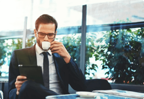 Man in pak op terras met een koffie in zijn hand en een ipad in zijn andere hand