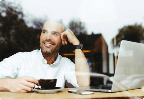 Man aan tafel bij het raam met koffie en zijn laptop kijkend naar buiten