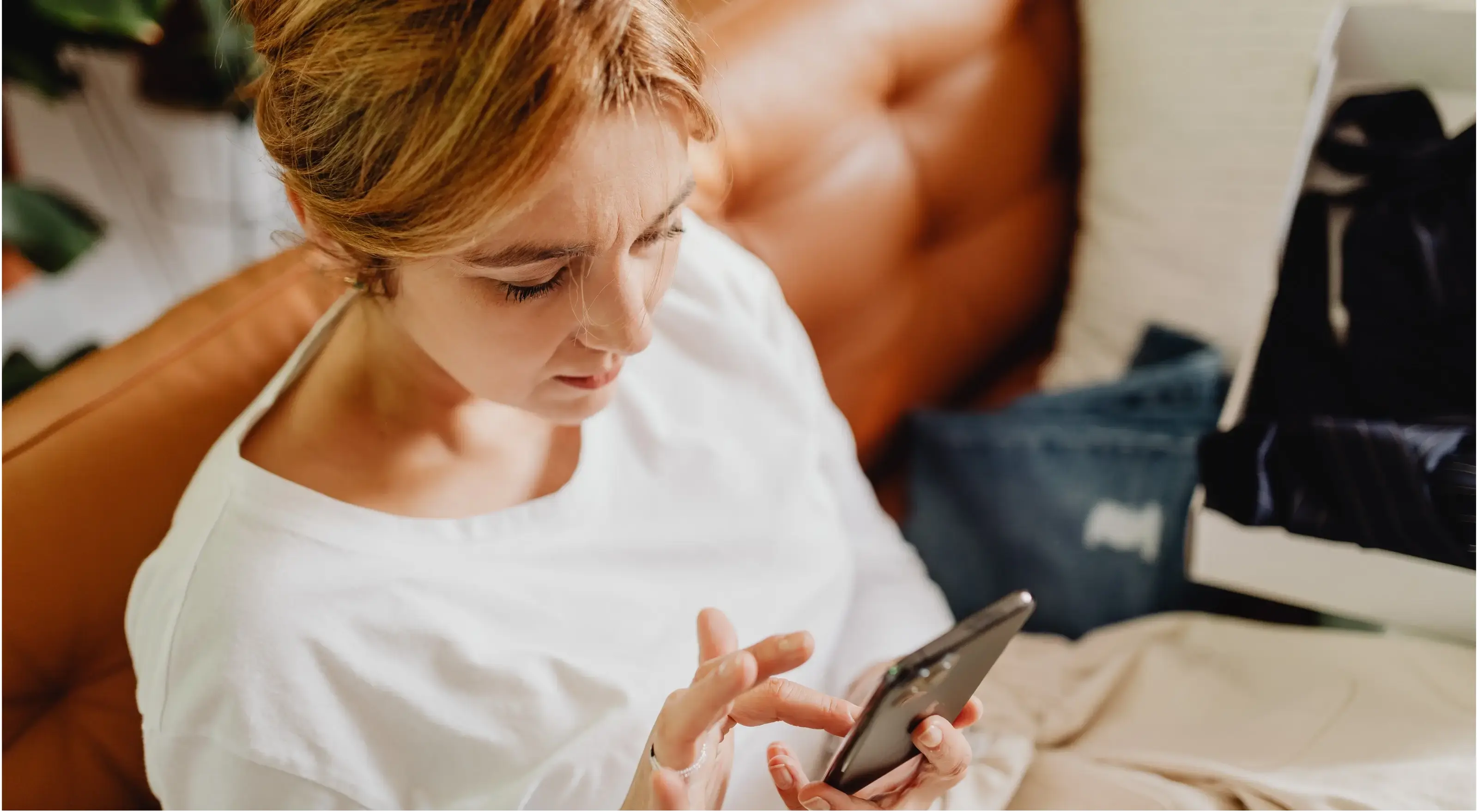 Vrouw in witte trui zit op een bruine bank en kijkt naar haar smartphone.